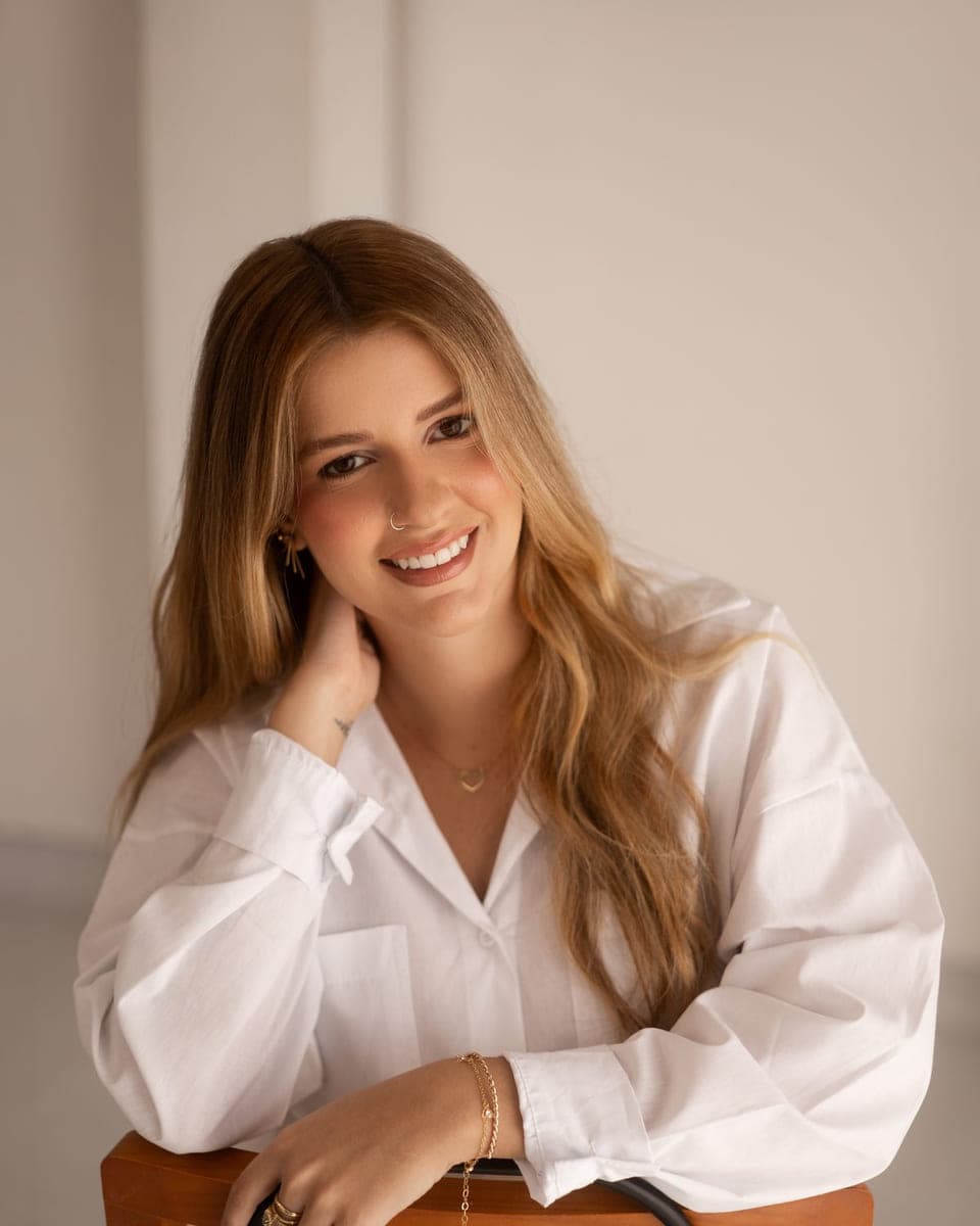 Retrato de Lara Pansiere, de camisa branca, apoiada no encosto de uma cadeira de madeira, sorrindo para a câmera.