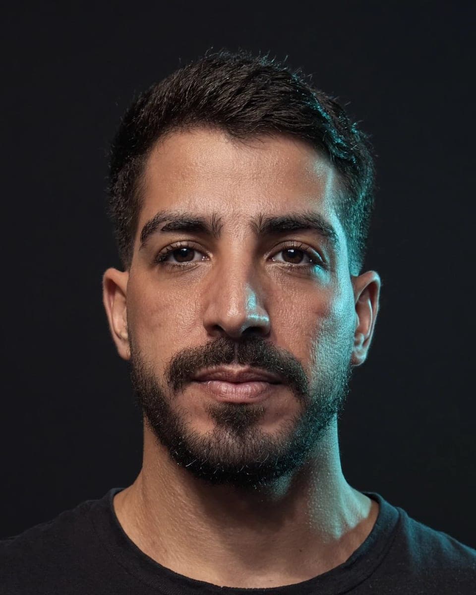 Retrato de Marcelo Souto, fundador do >STO_, de cabelo curto, barba e camiseta preta, sobre fundo escuro, olhando para a câmera.